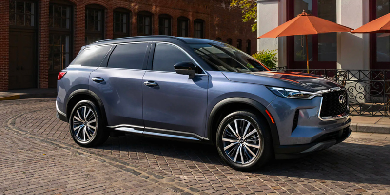 A blue 2025 INFINITI QX60 SUV parked on a cobblestone street next to a building with large windows.