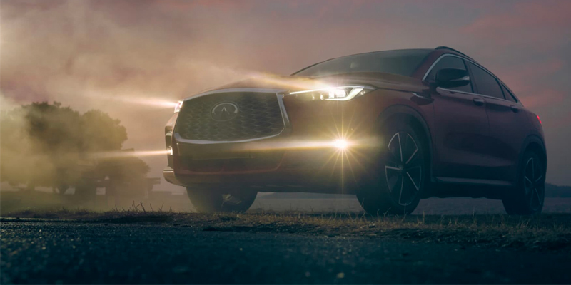 A red 2025 INFINITI QX55 SUV parked on a road surrounded by fog, with its headlights on.