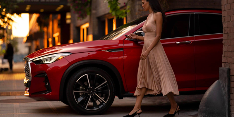 A woman in a beige dress walks by a red 2025 INFINITI QX55 parked on a city street at sunset.