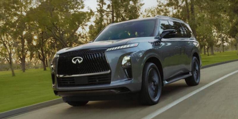 Dark grey INFINITI QX80 luxury SUV driving on a tree-lined road.