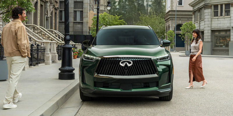 A green 2026 INFINITI QX60 Autograph is parked on a city street between two pedestrians, highlighting its bold front grille, premium design, and upscale urban styling.